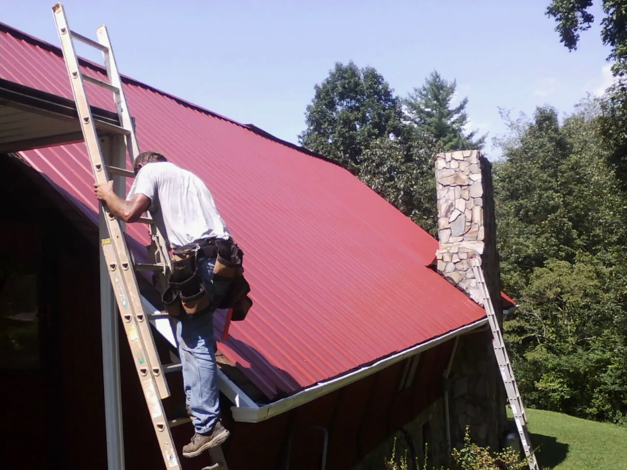 Roofer on a ladder beside a red metal roof and stone chimney — Soffit & Fascia Repair by Roofing Brothers in Washburn
